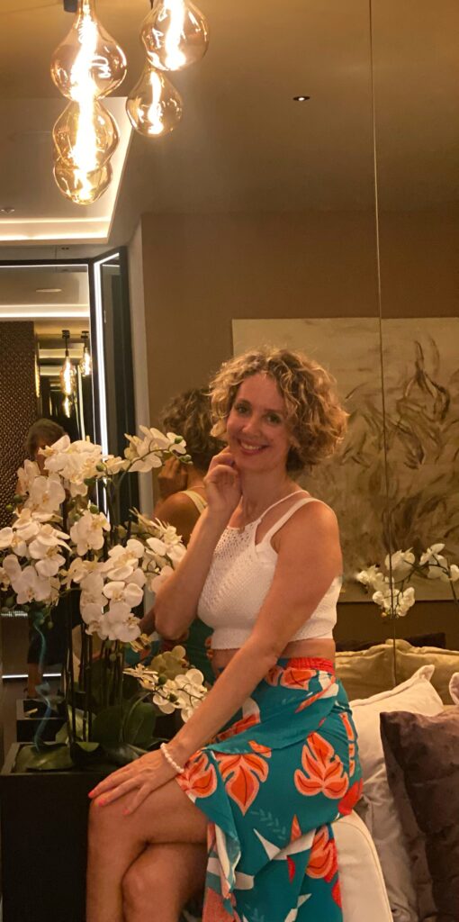 Dr. Carolina Díez Jorge Psicóloga posando entre flores orquídeas vestida con una falda muy luminosa de flores naranjas en fondo verde. Esta sonriendo 