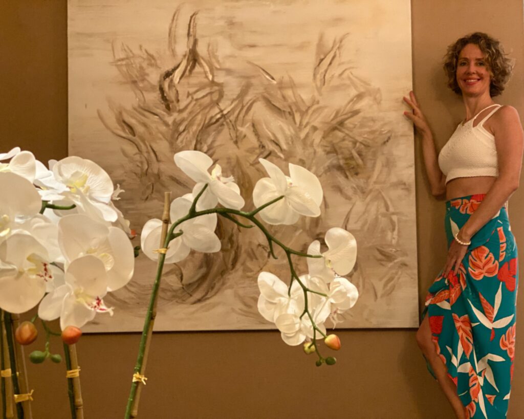 Mujer posando junto a una pintura al oleo con falda de colores, sonriente y de fondo unas flores orquídeas
